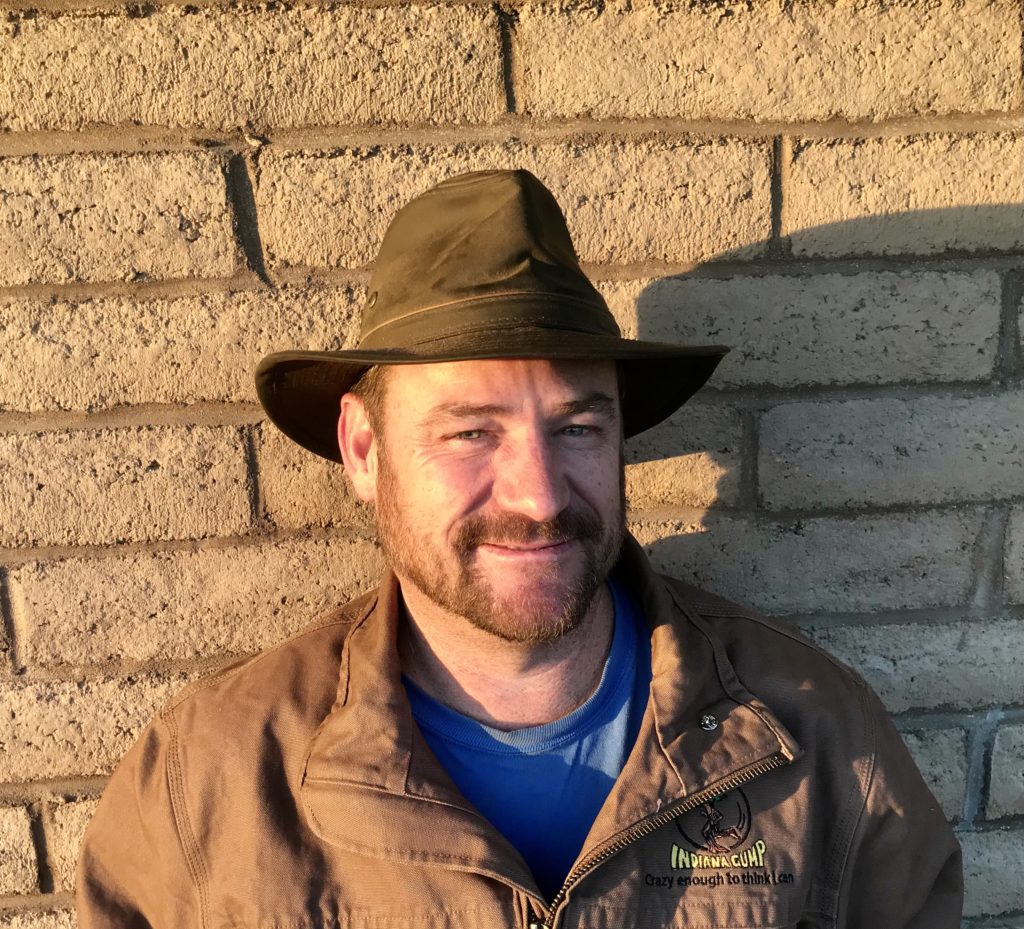 photo of man with short beard in a fedora and brown jacket standing in front of a brick wall